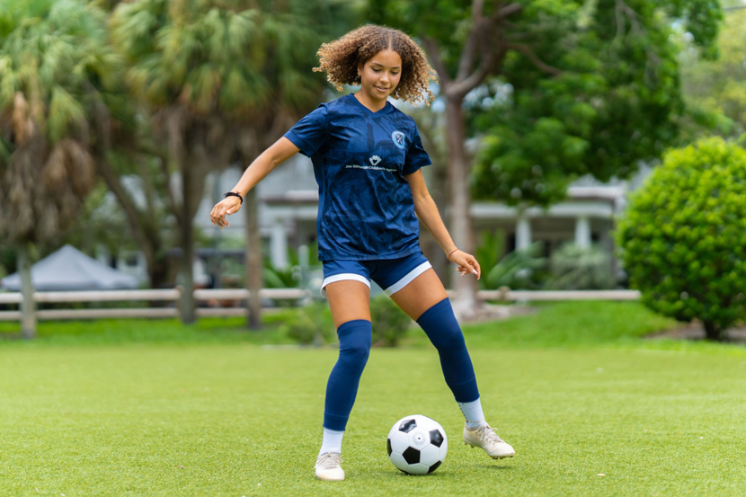 Young girl wearing blue jersey holding soccer ball in front of soccer field.